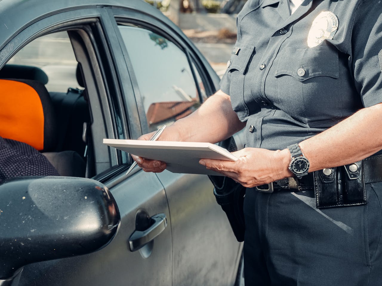 A police officer in uniform writes a ticket to a driver through the car window on a sunny day.